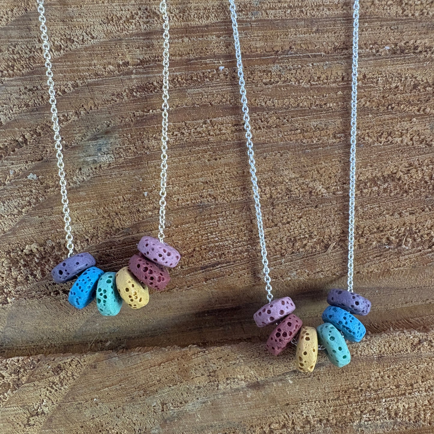 Two chain necklaces with clusters of small lava, rondel beads in pastel purple, blue, yellow, green, and pink displayed on a rustic wooden surface.