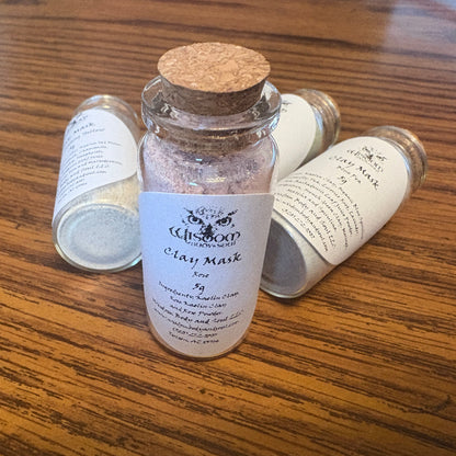 Three small glass jars of Clay Mask and skin scrub by Wisdom Body and Soul, one standing upright in the center and two lying on their sides behind it, all on a wooden tabletop.