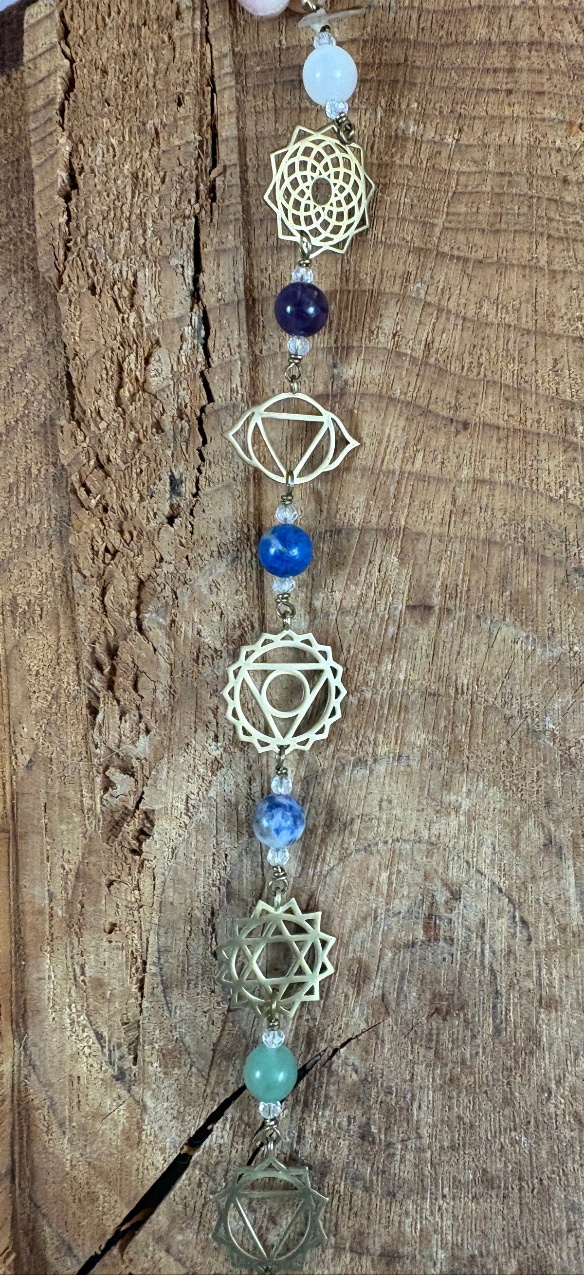 A beaded Chakra wall hanging featuring geometric chakra metal charms. Red, orange, yellow, green, blue, purple gemstones wire wrapped.  Displayed vertically on a rustic wood slice background