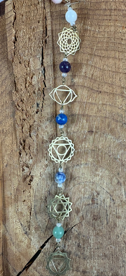 A beaded Chakra wall hanging featuring geometric chakra metal charms. Red, orange, yellow, green, blue, purple gemstones wire wrapped.  Displayed vertically on a rustic wood slice background