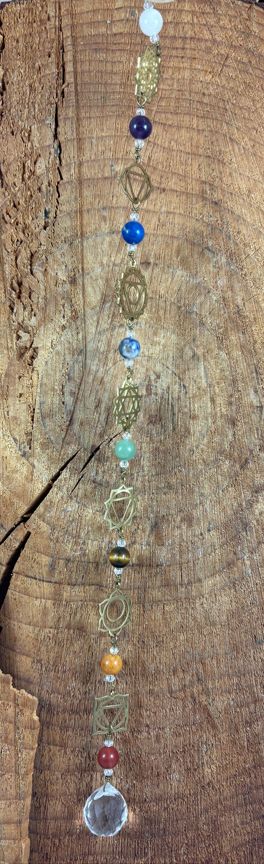 A beaded Chakra wall hanging featuring geometric chakra metal charms. Red, orange, yellow, green, blue, purple gemstones wire wrapped.  Displayed vertically on a rustic wood slice background
