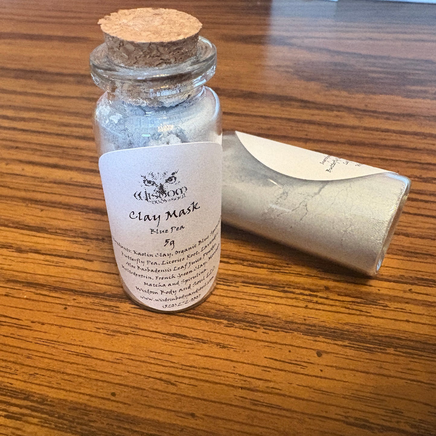 A small glass vial with a cork stopper and a white label reading Clay Mask  shown on a wooden table next to a jar of the same mask.