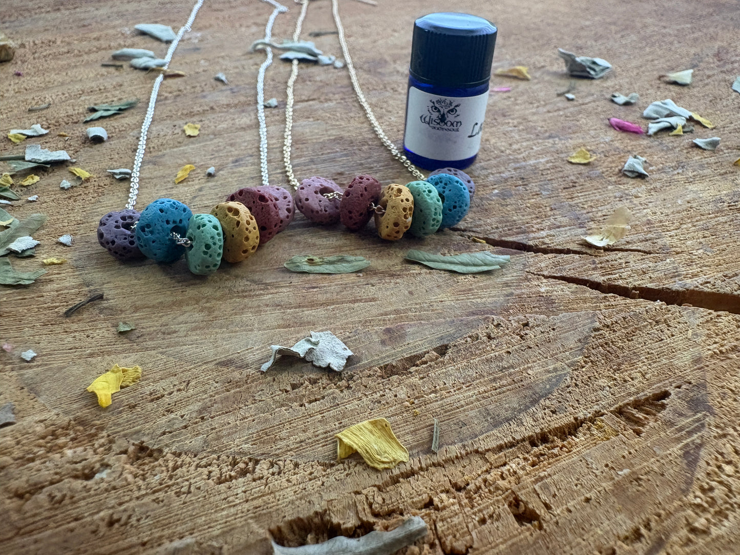 A small bottle of essential oil with a white label, dark blue cap, and black lid, placed on a rustic wooden surface next two chain necklaces of colorful rondel lava beads.