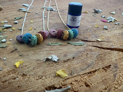 A small bottle of essential oil with a white label, dark blue cap, and black lid, placed on a rustic wooden surface next two chain necklaces of colorful rondel lava beads.