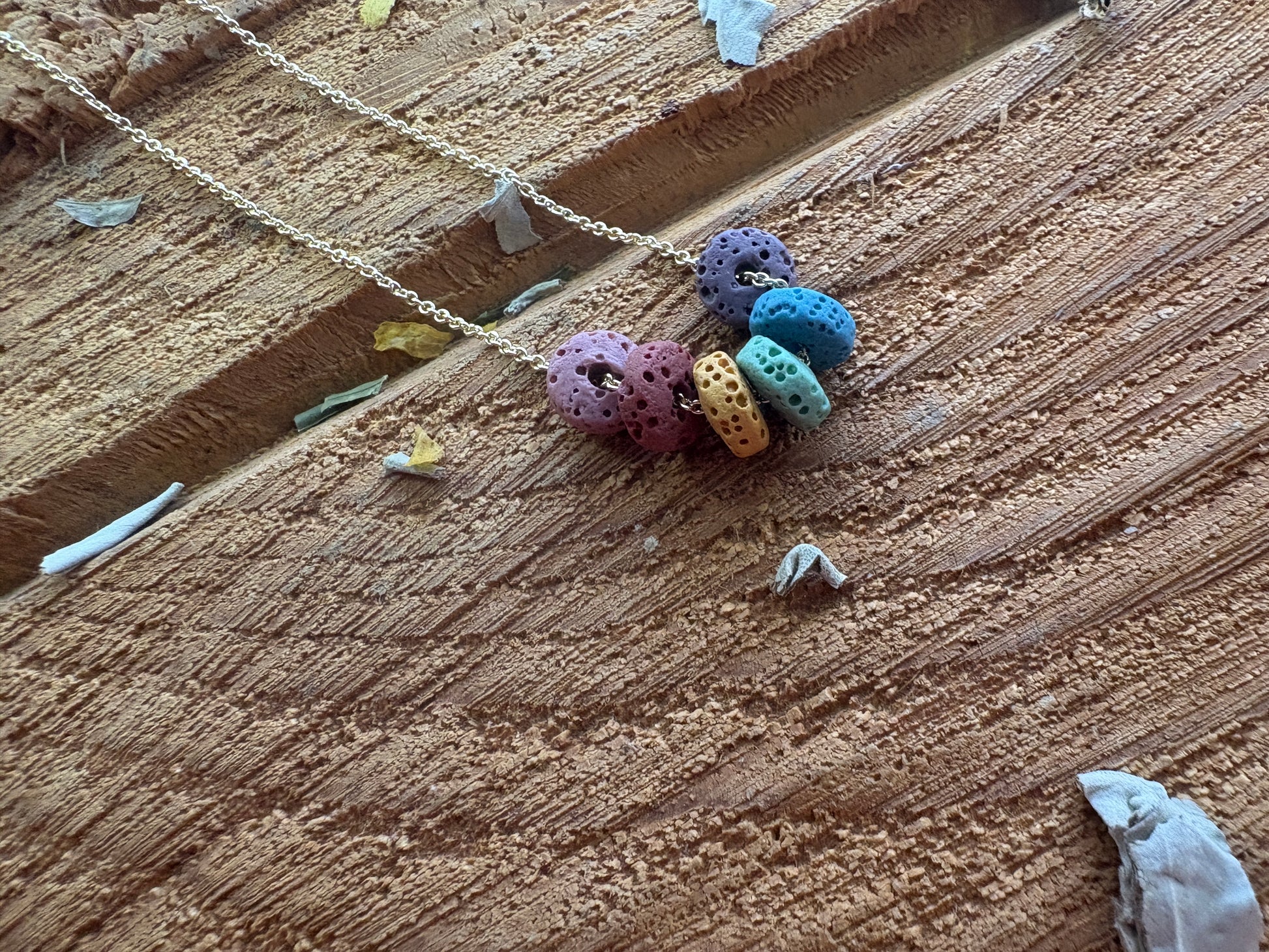 Necklace with five porous lava beads. Colorful lava beads in purple, blue, yellow, green and pink on a gold chain, laid on a textured wooden surface.
