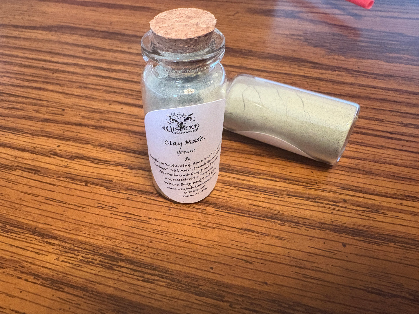A small glass jar of Clay Mask Green clay mask powder with a cork stopper and a white label, shown next to a rolled-up jar of the same product on a wooden table.