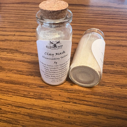 Two small glass jars containing clay mask powder on a wooden table. One jar is upright with a cork stopper and a white label reading 'CLAY MASK Illuminating Yellow 5g', and the other is lying on its side showing the powder inside.