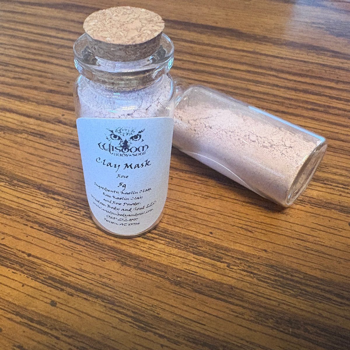 A small glass jar with a cork stopper and a white label reading 'Clay Mark' sits on a wooden table, with a second jar lying beside it showing the loose clay powder inside.