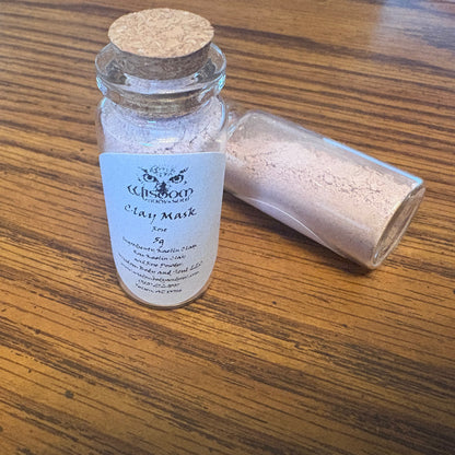 A small glass jar with a cork stopper and a white label reading 'Clay Mark' sits on a wooden table, with a second jar lying beside it showing the loose clay powder inside.