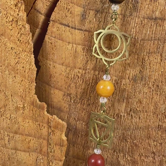 A beaded Chakra wall hanging featuring geometric chakra metal charms. Red, orange, yellow, green, blue, purple gemstones wire wrapped.  Displayed vertically on a rustic wood slice background