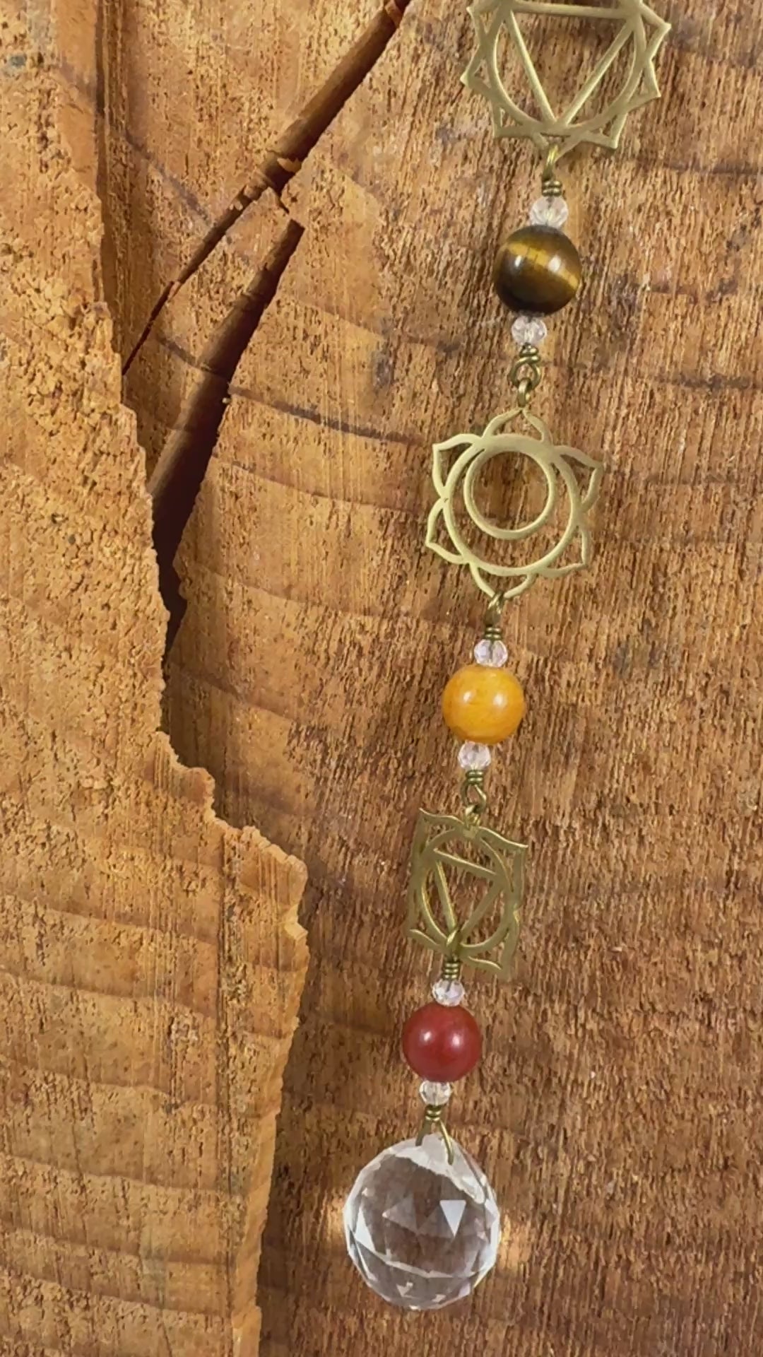 A beaded Chakra wall hanging featuring geometric chakra metal charms. Red, orange, yellow, green, blue, purple gemstones wire wrapped.  Displayed vertically on a rustic wood slice background
